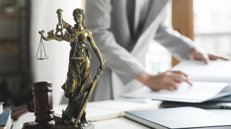 A lawyer is reviewing documents on a desk behind a gavel and a Lady Justice sculpture